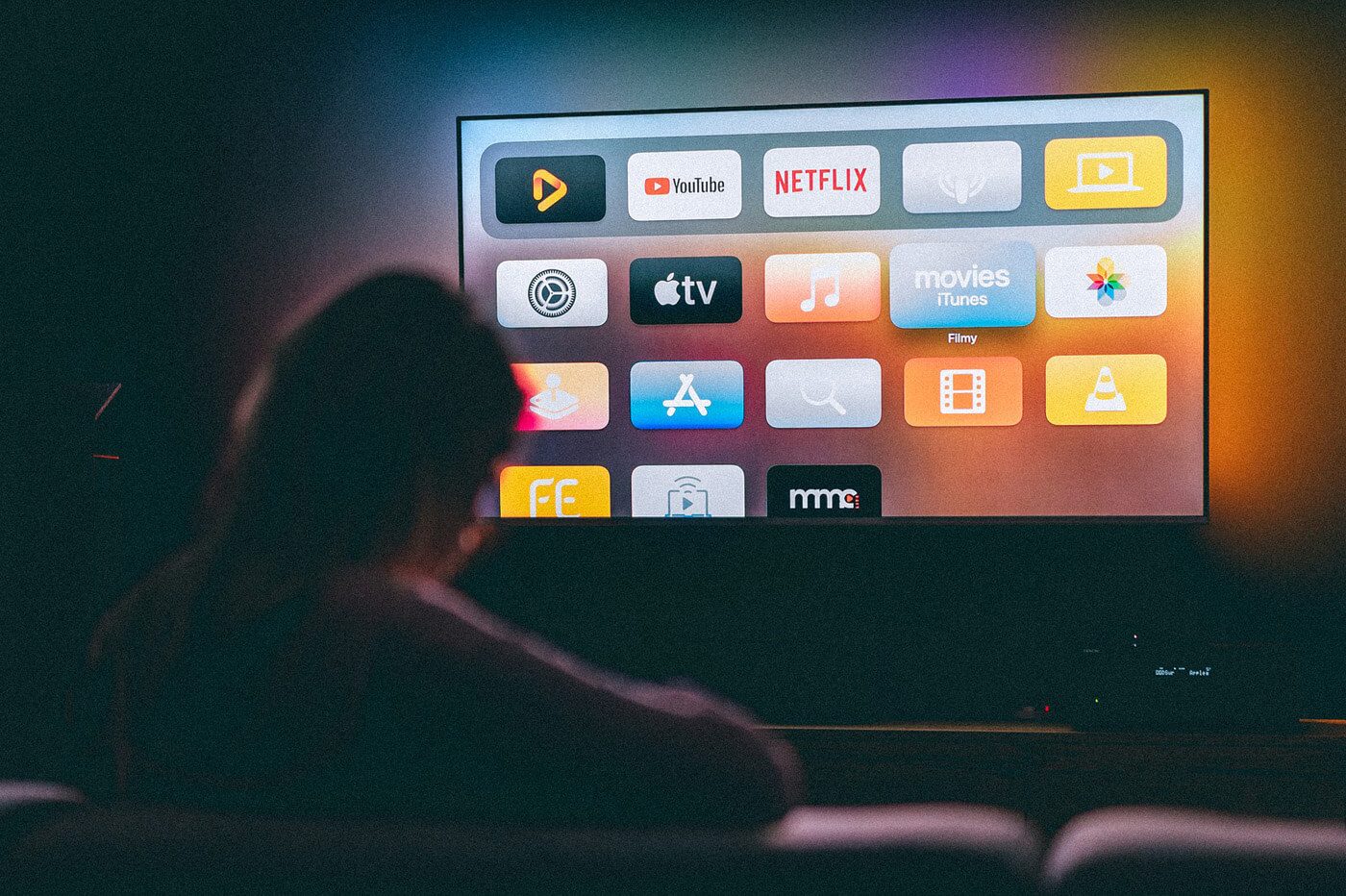 A person sits in a dimly lit room, facing a large screen displaying various app icons including YouTube, Netflix, Apple TV, and others. The room has a dark ambiance, and the screen glows brightly with vibrant colors from the app icons.